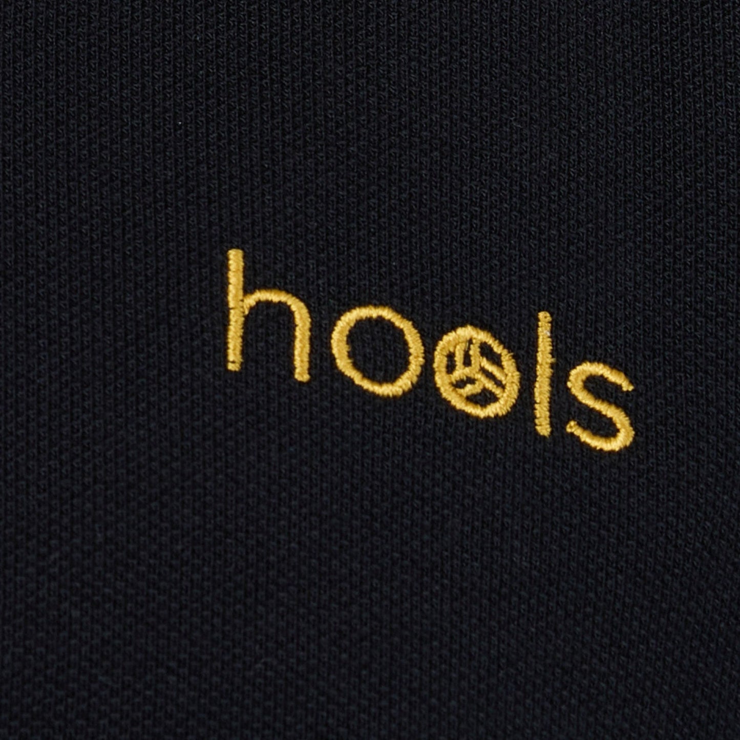 Detalle del logo bordado Hools en polo negro con rayas amarillas