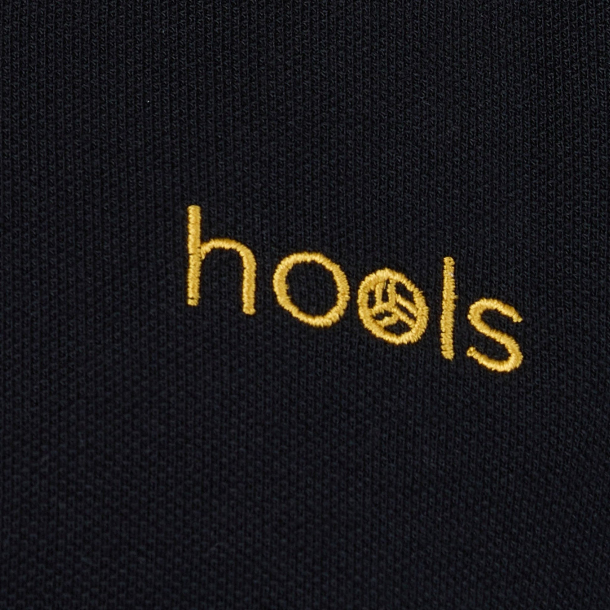 Detalle del logo bordado Hools en polo negro con rayas amarillas